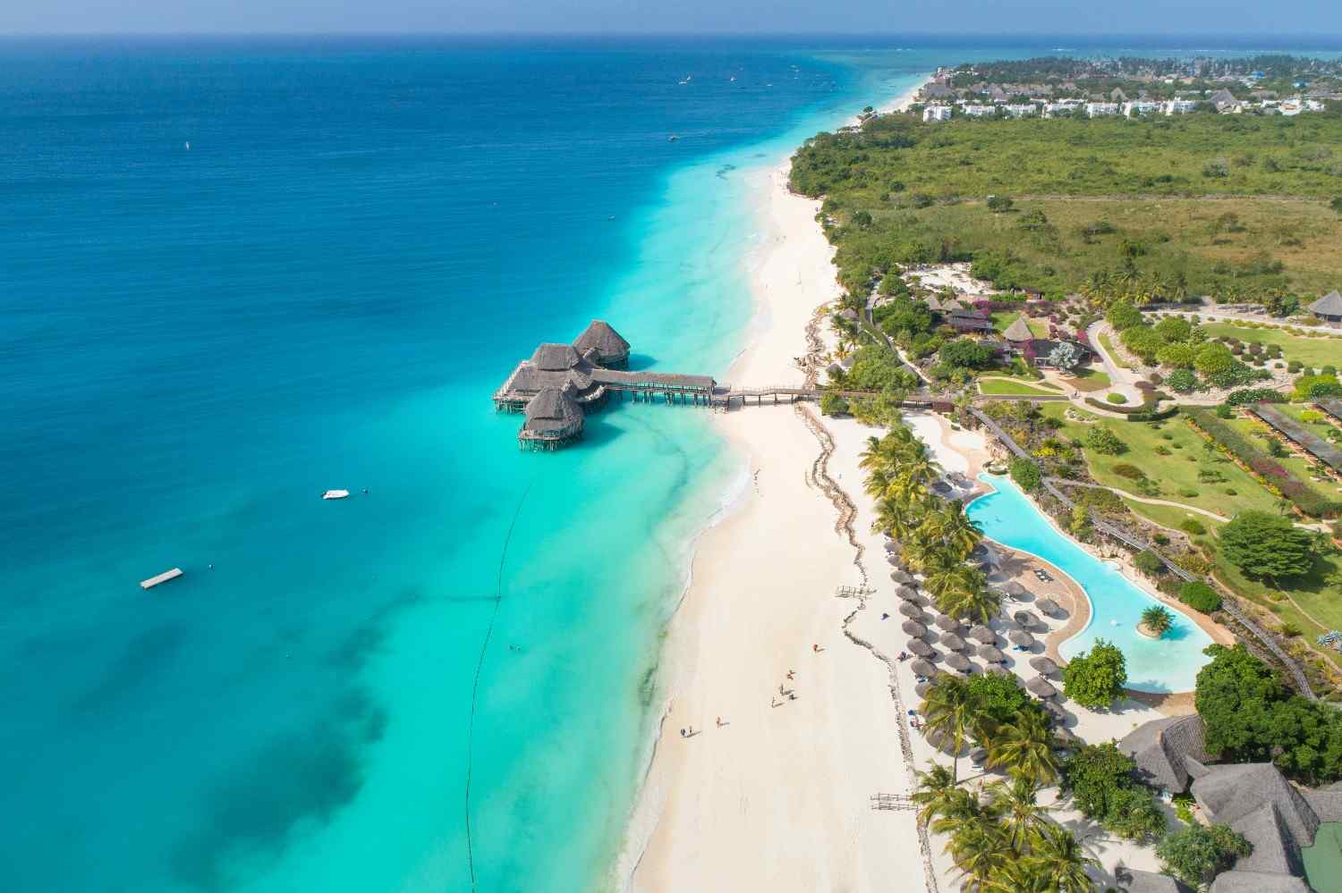 Widok z lotu ptaka na rajską plażę Nungwi na Zanzibarze. Nad turkusową wodą Oceanu Indyjskiego unosi się drewniane molo z restauracją krytą strzechą, a obok rozciąga się biała, piaszczysta plaża i luksusowy resort z basenem otoczonym palmami.